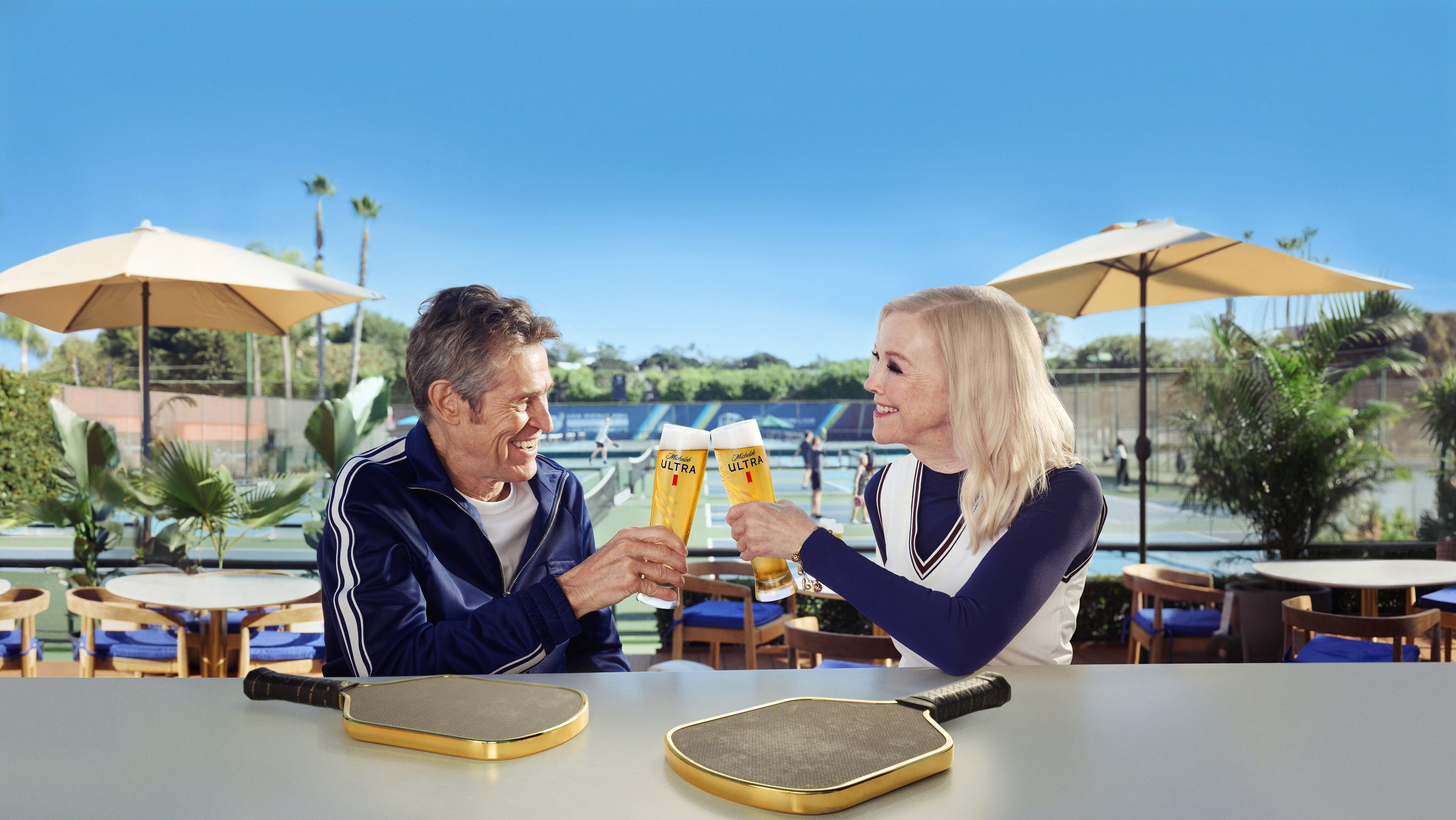 Willem Dafoe and Catherine O'Hara holding High Roller chrome-gold pickleball paddles in a Michelob ULTRA campaign scene