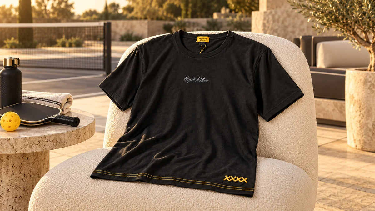 The High Roller Lounge T-shirt draped over a cream courtside chair, showing tonal script and gold stitch details.