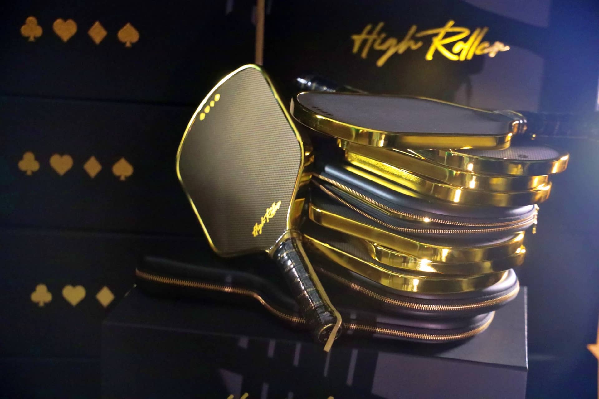 High Roller black pickleball paddle with a chrome gold edge beside stacked matching paddle cases