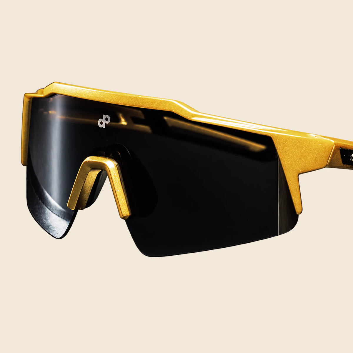 Close-up of High Roller sunglasses with their continuous black shield lens, angular gold frame and small white DP mark on warm cream.