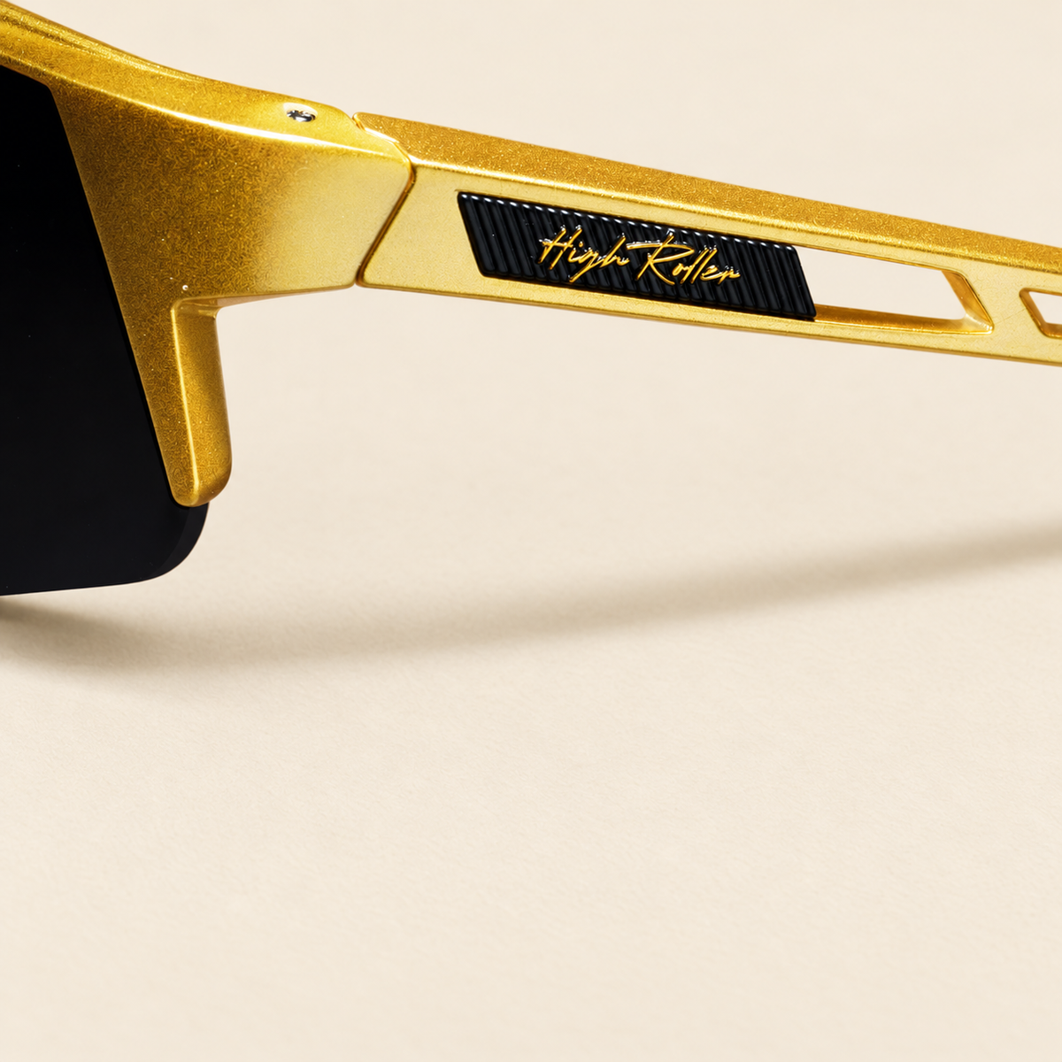 High Roller sunglasses temple with gold script on a ribbed black inset and gold frame, shown on cream.