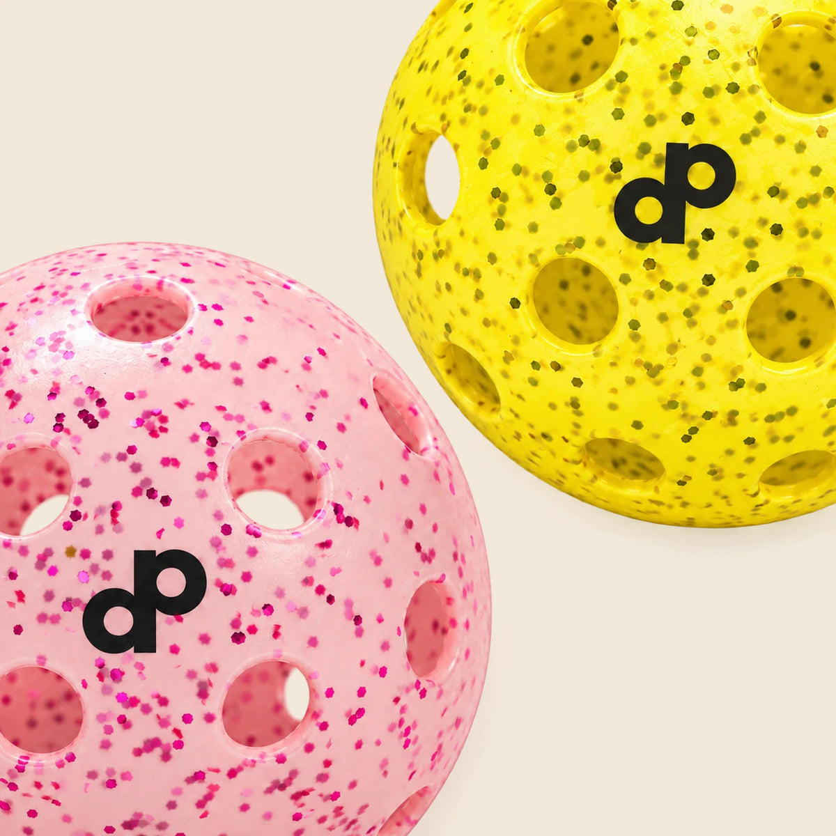 Cropped color and material detail of pink and yellow glitter pickleballs side by side on warm cream.