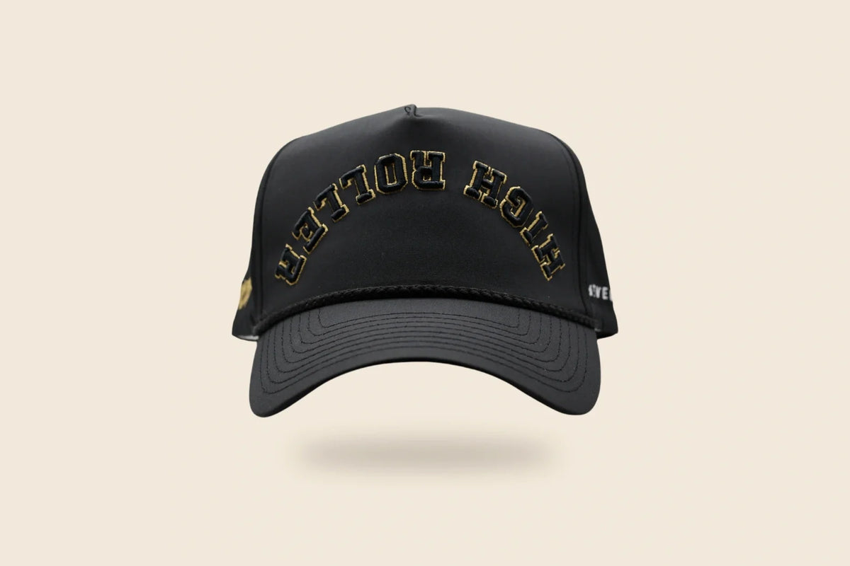 Black High Roller hat with reversed lettering, gold embroidery trim and a soft floating shadow on an off-cream background.