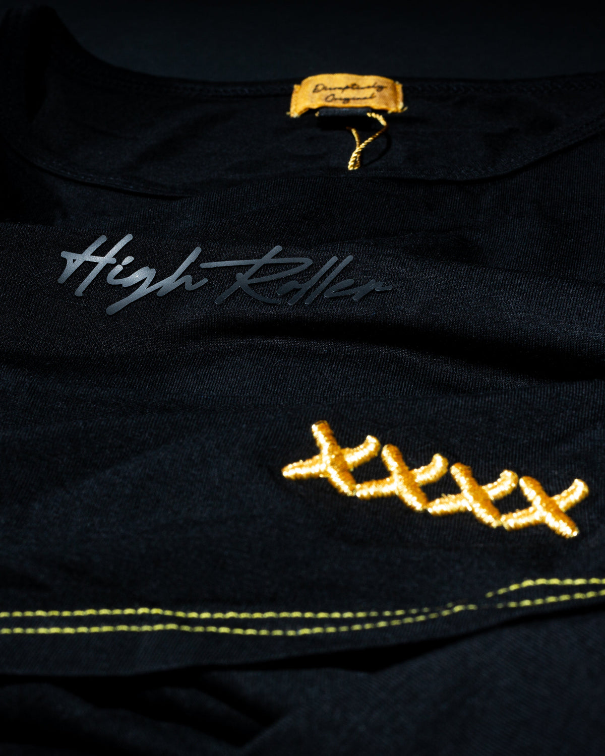 Black High Roller shirt showing the script print, four curved gold X marks and gold hem stitching.