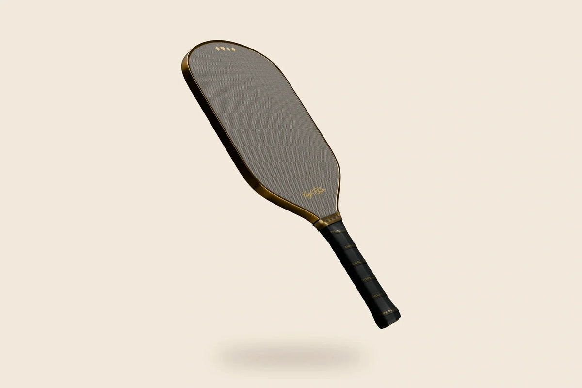 High Roller paddle with reflective gold perimeter, patterned face, suit-symbol band and original grip lettering, floating on an off-cream background.