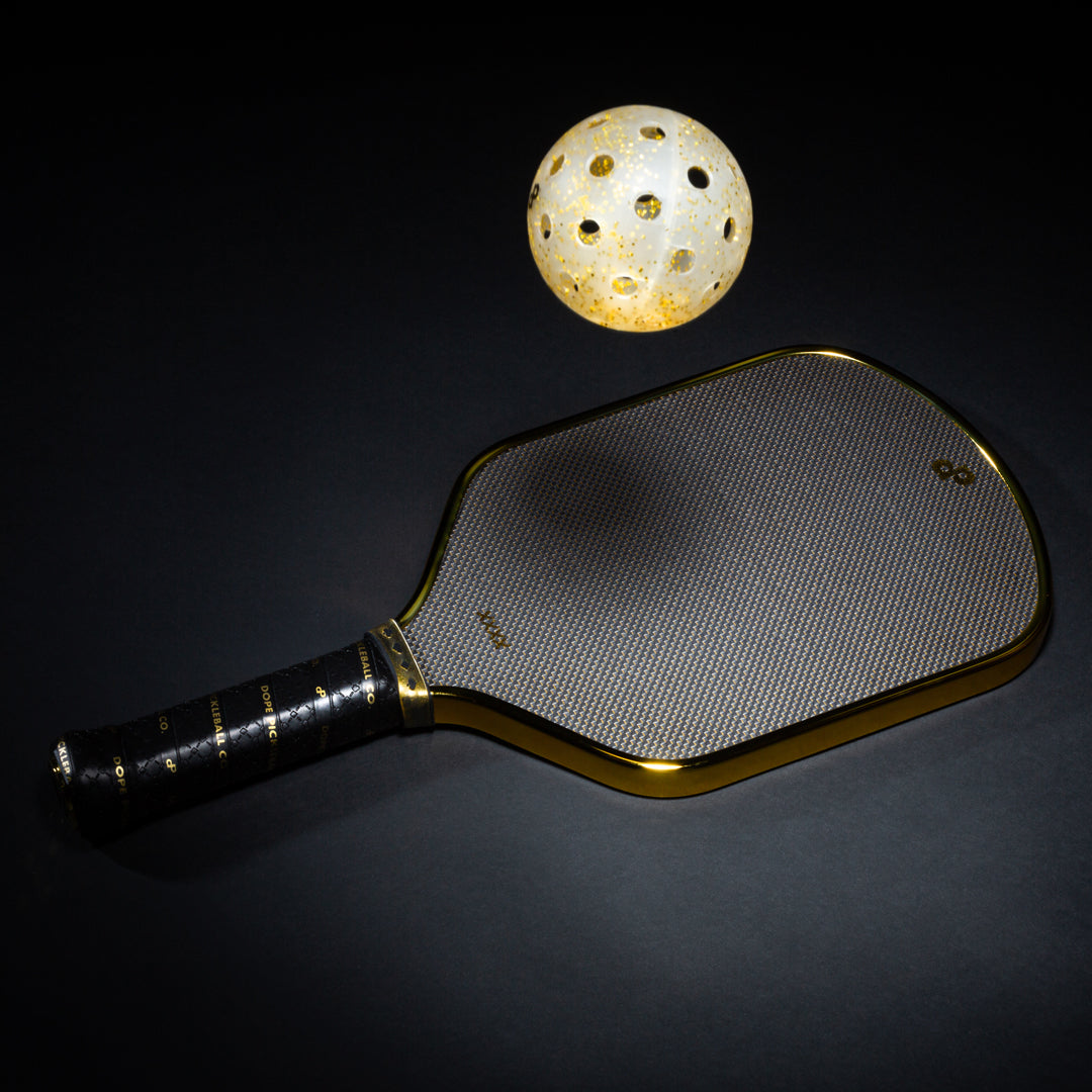 High Roller black pickleball paddle with a chrome-gold edge beside a gold glitter pickleball