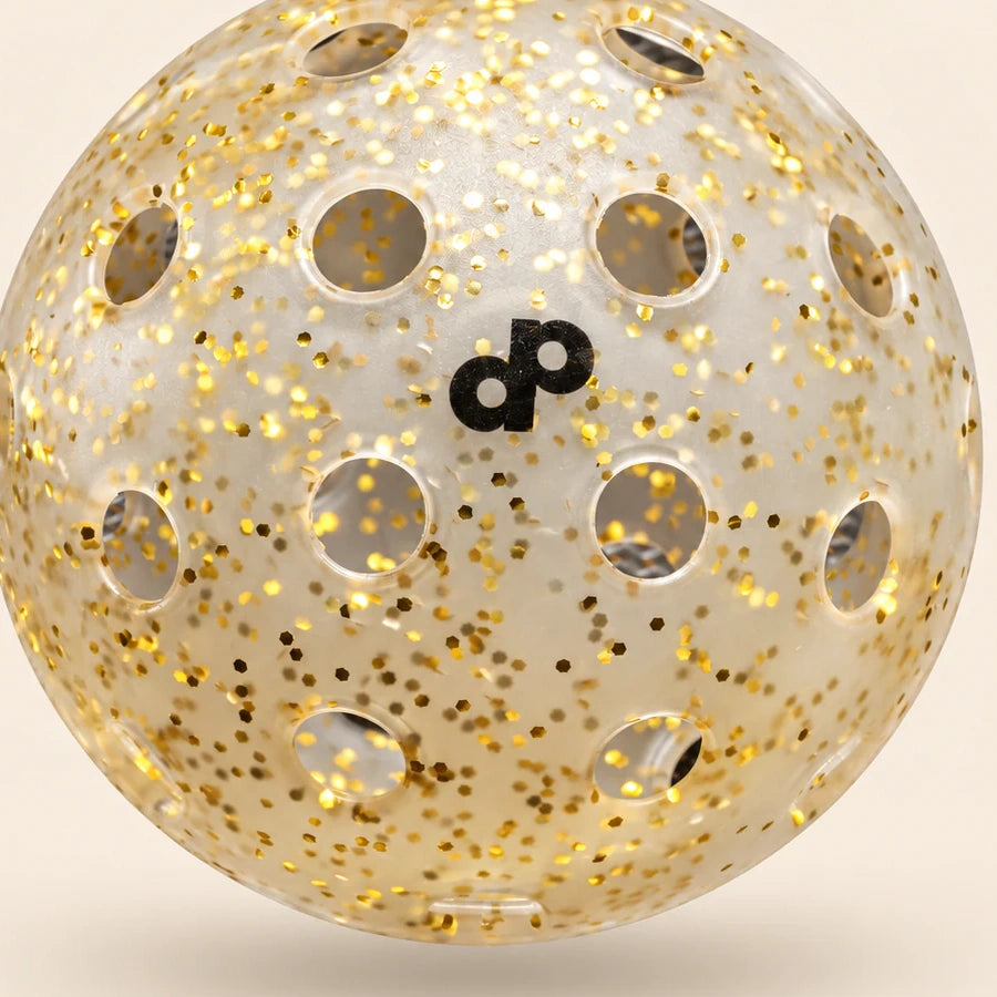 Close-up of the clear High Roller gold-glitter pickleball, black dp print, embedded flakes and circular holes.