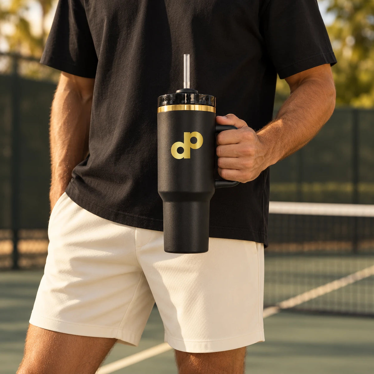 Black and gold High Roller tumbler held by its handle courtside, styled with a black T-shirt and ivory shorts.