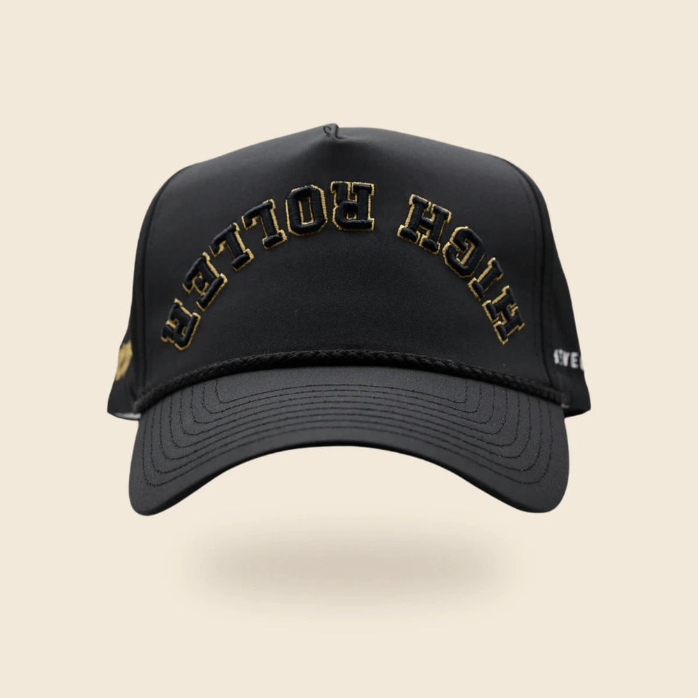 Black High Roller reversed-lettering hat with gold embroidery trim, floating above an off-cream background and soft shadow.