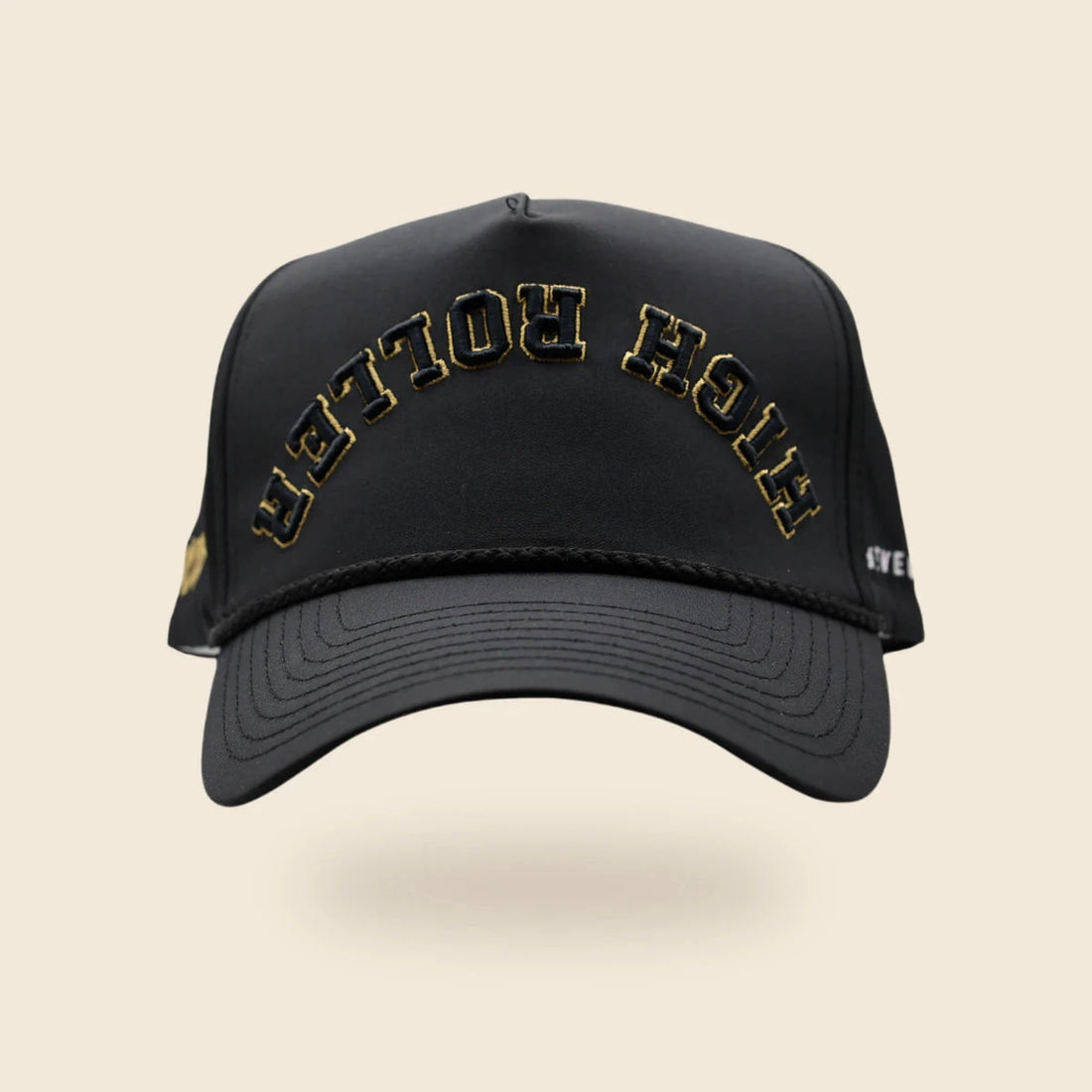 Black High Roller reversed-lettering hat with gold embroidery trim, floating above an off-cream background and soft shadow.