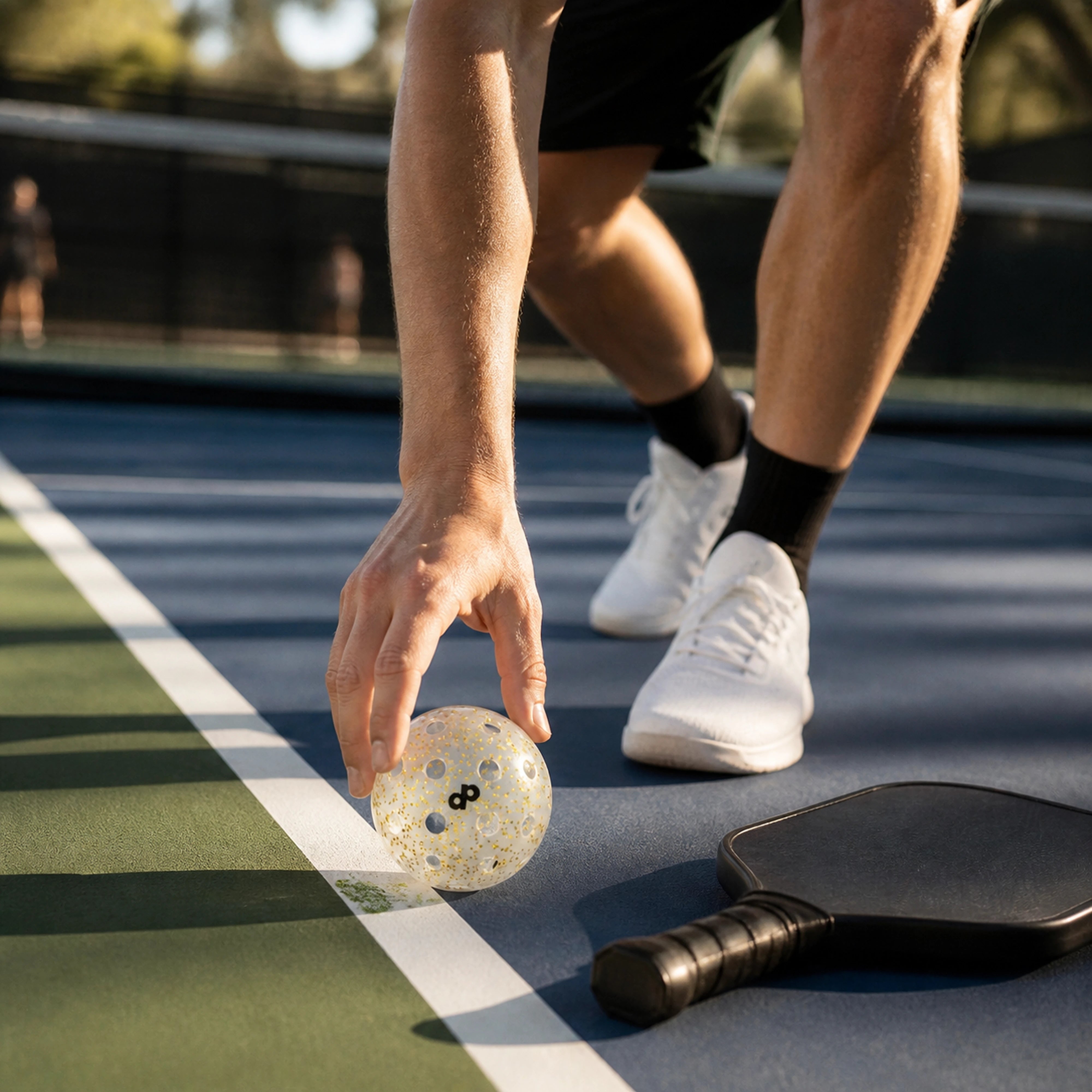 Player picking up a clear gold glitter DOPE pickleball beside a paddle on an outdoor court