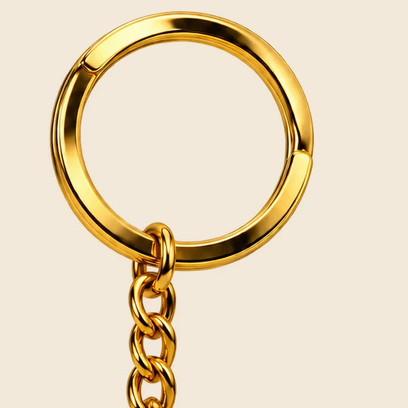 Close-up of the gold logo keychain split ring, chain attachment and polished gold-tone finish on cream.