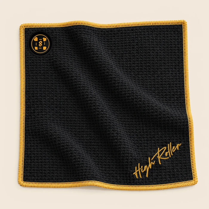 Black High Roller towel with gold edges and embroidered script; the fixed DP magnet aligns with the upper-left corner.