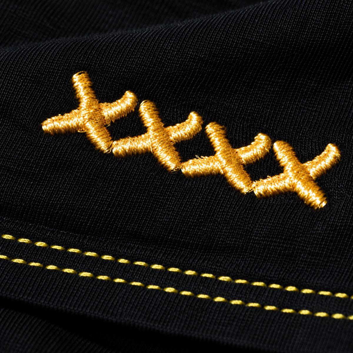 Micro photograph of the High Roller shirt’s linked gold XXXX embroidery and two fine gold hem stitching rows on black jersey.