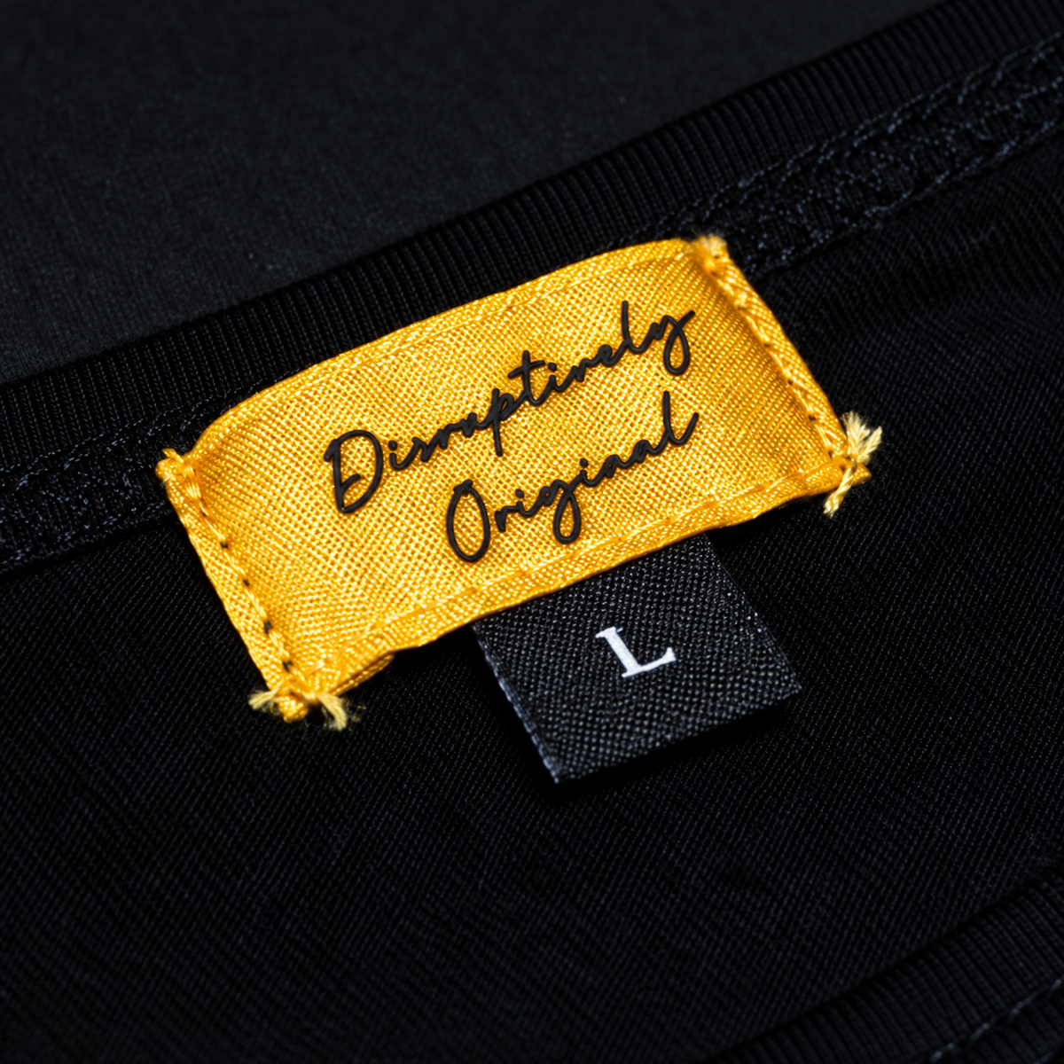 Close-up of the sewn gold Disruptively Original shirt label with black rubber script and the black L size tab.