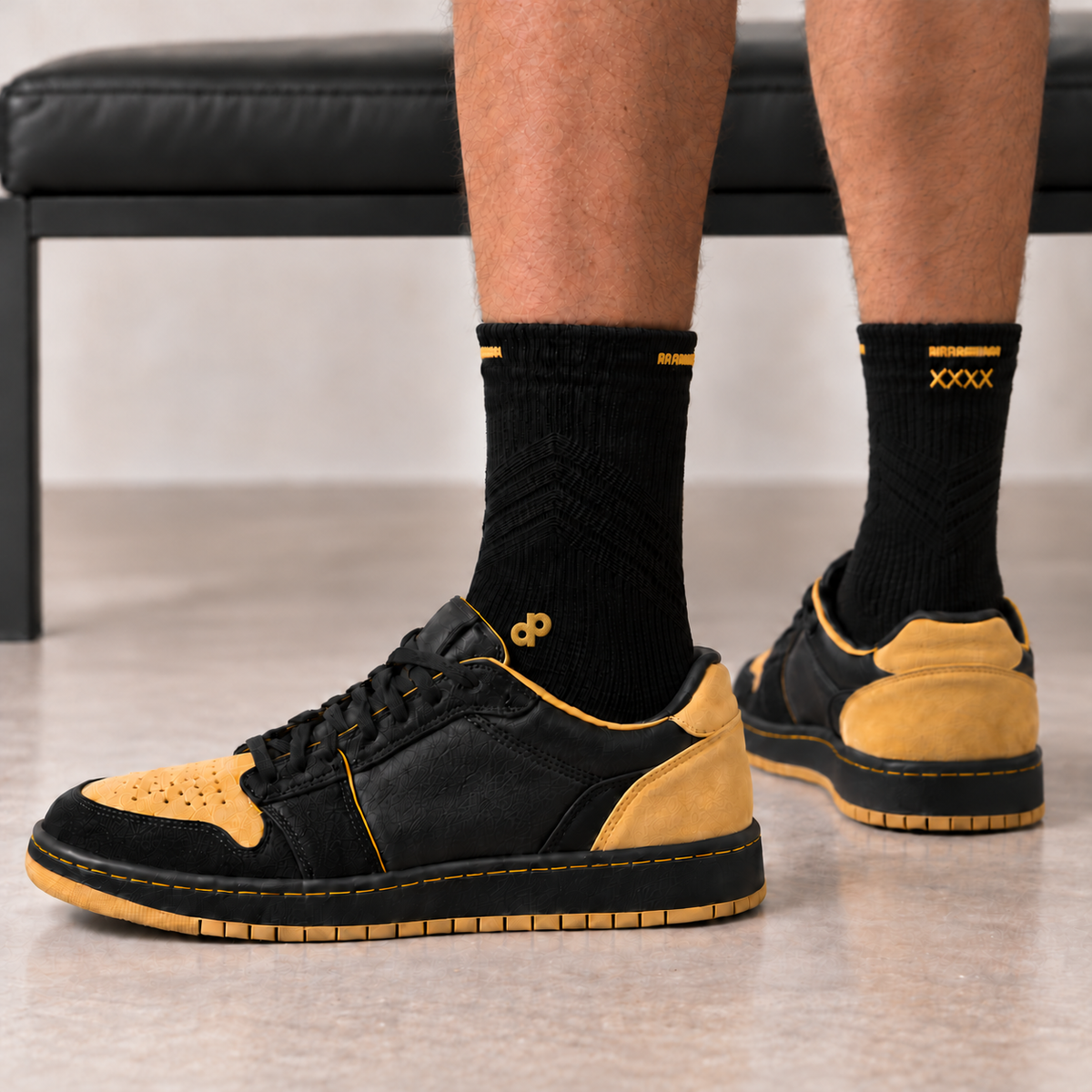 High Roller black socks worn with unbranded black and gold low-top shoes, with small connected gold DP embroidery at the outside-front ankle, separated front and rear cuff lines, and small rear XXXX stitching.