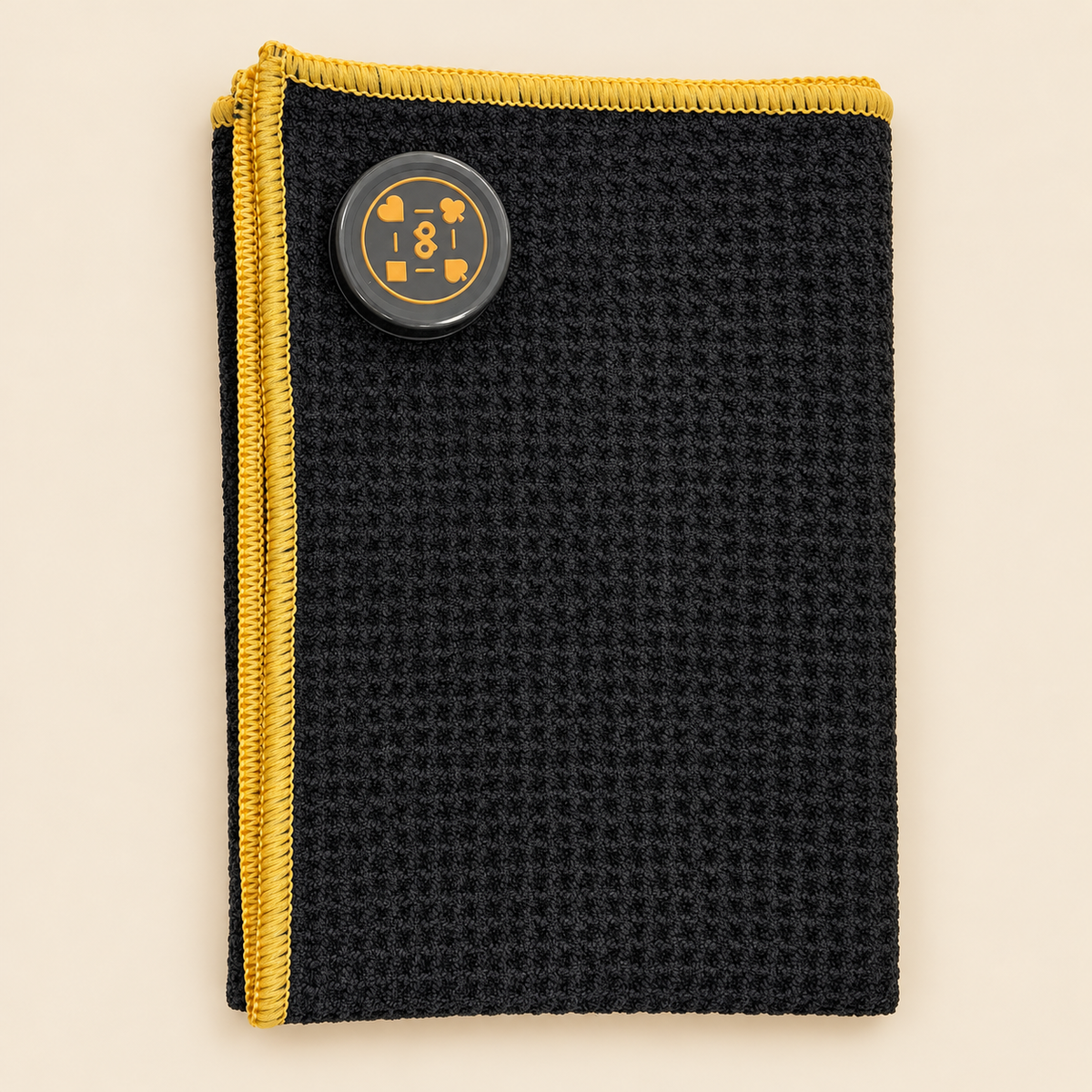 Folded High Roller towel lying flat on a cream tabletop, with its original sewn magnet, fine black fabric and gold overlock stitching.