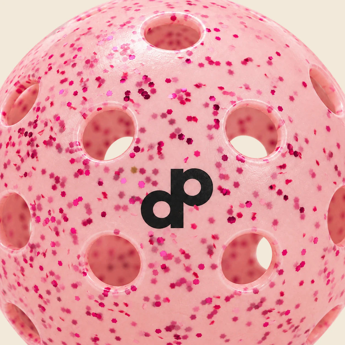 Close-up of the pink glitter pickleball's black dp logo, embedded pink flakes and surrounding holes on cream.