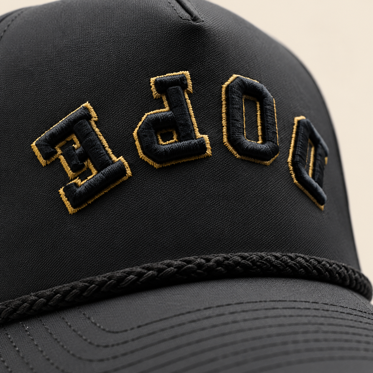 Close view of the black DOPE hat with reversed black embroidery, gold thread outlines and a black braided cord.