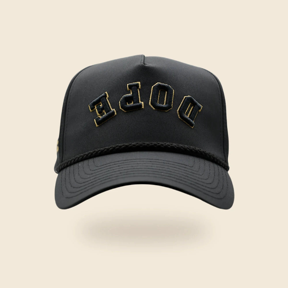 Black Dope reversed-lettering hat with gold embroidery trim, floating above a soft shadow on an off-cream background.