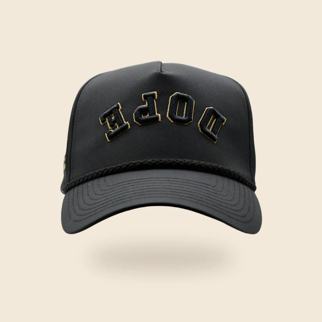 Black DOPE reversed-lettering hat with gold embroidery trim, floating above a soft shadow on an off-cream background.