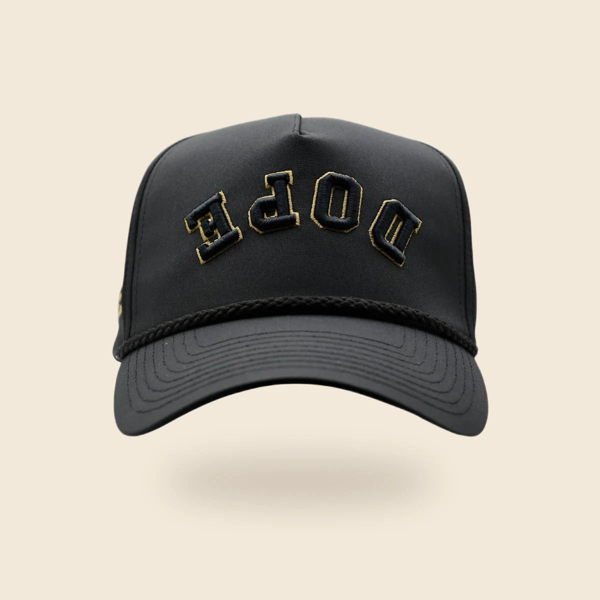 Black DOPE reversed-lettering hat with gold embroidery trim, floating above a soft shadow on an off-cream background.