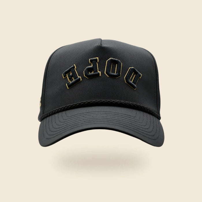 Black DOPE reversed-lettering hat with gold embroidery trim, floating above a soft shadow on an off-cream background.