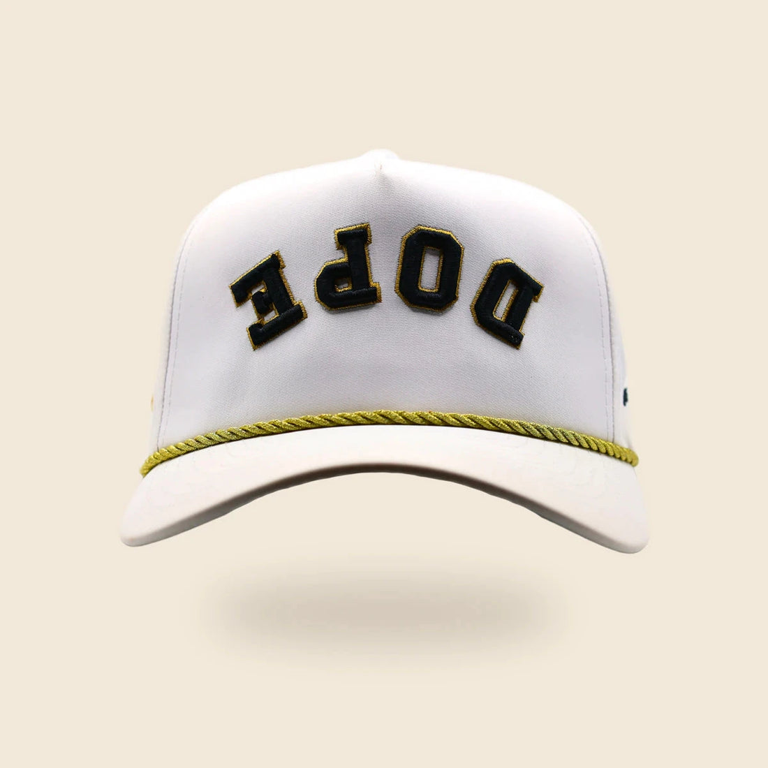 White DOPE reversed-lettering hat with black-and-gold embroidery and gold cord, floating over an off-cream background.