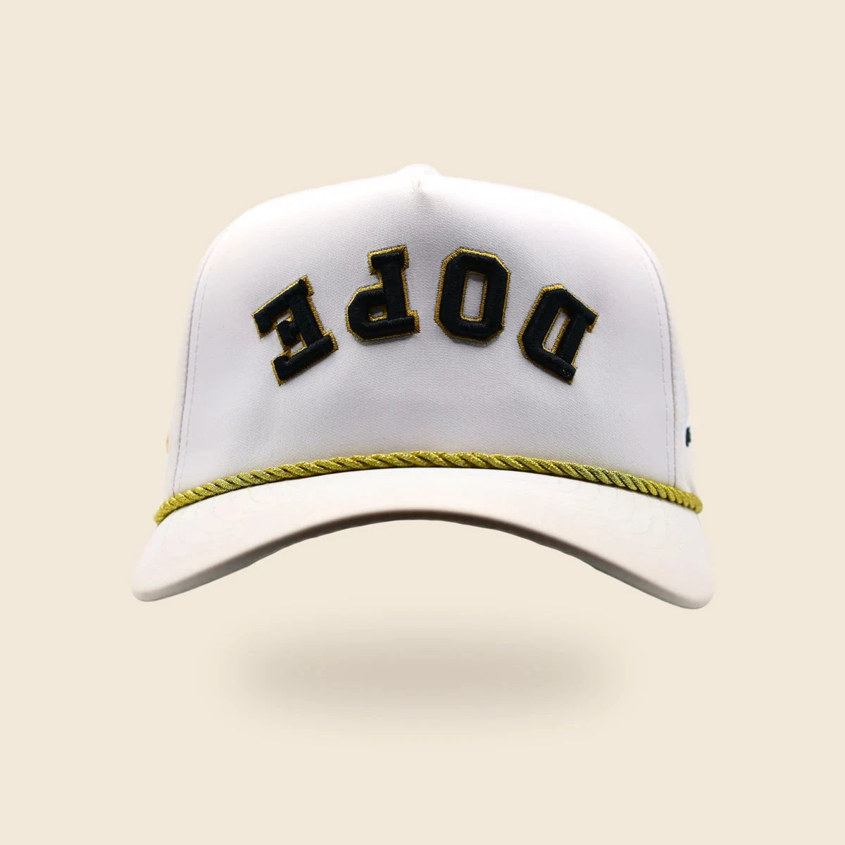 White DOPE reversed-lettering hat with black-and-gold embroidery and gold cord, floating over an off-cream background.