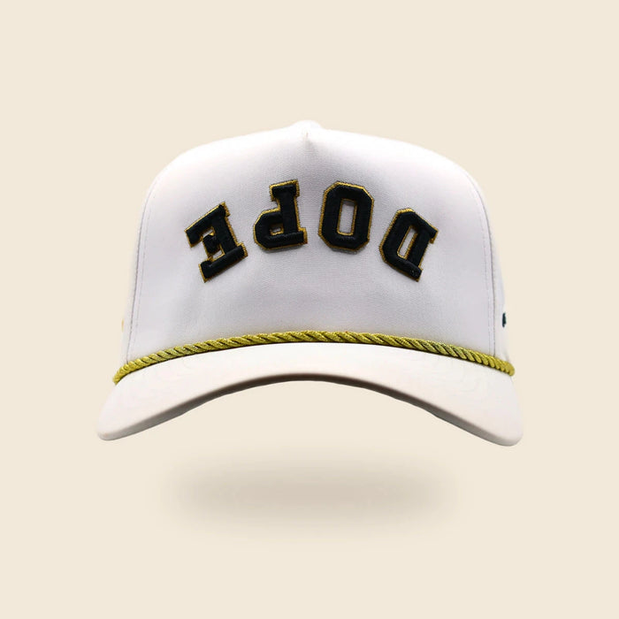 White DOPE reversed-lettering hat with black-and-gold embroidery and gold cord, floating over an off-cream background.