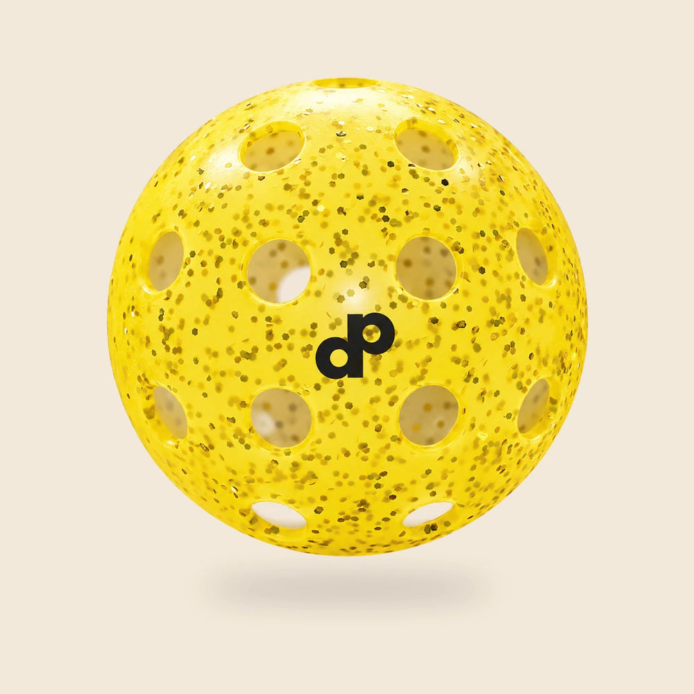 Single yellow Dope glitter pickleball with black DP logo and circular holes, floating on cream.