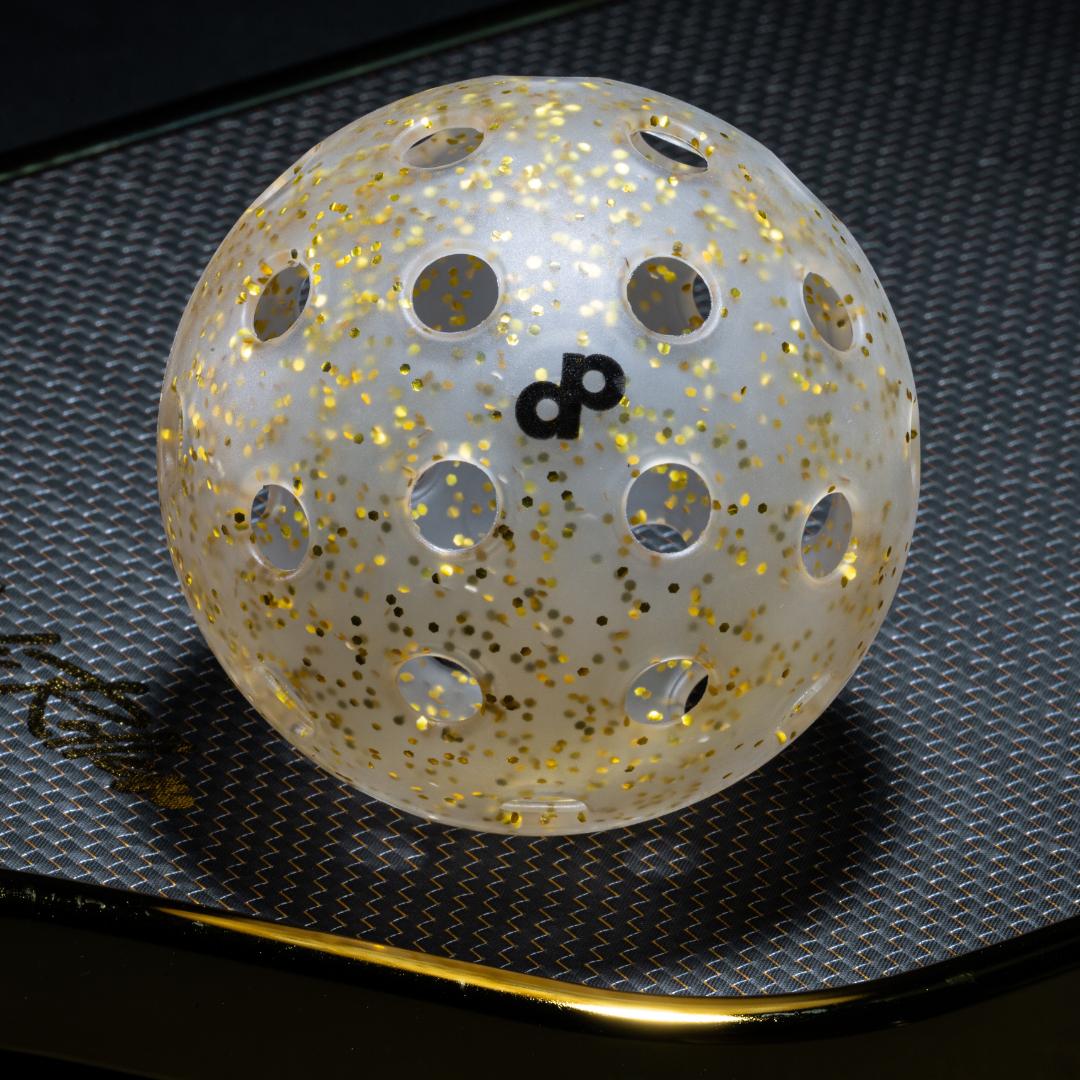 A closeup photo of the High Roller Gold Glitter-Infused translucent pickleball ball by Dope Pickleball Co resting on the 3K Carbon Weave surface of the High Roller Paddle.