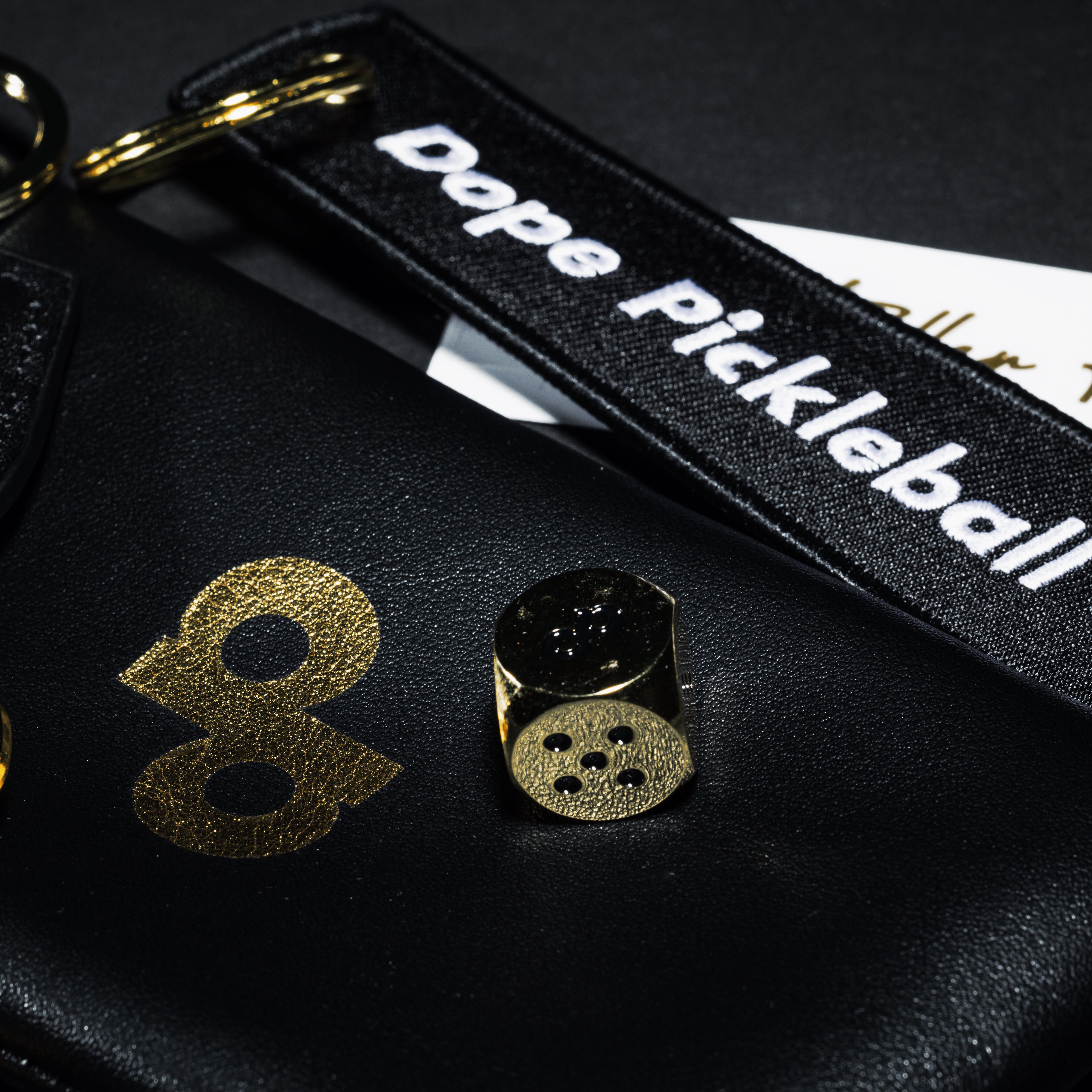 Black Dope Pickleball strap, gold dp logo on a black pouch, and black-and-gold die