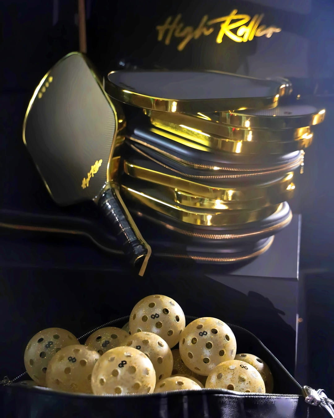 A luxury gym bag full of glittery pickleballs in front of a stack of gold rimmed pickleball paddles.