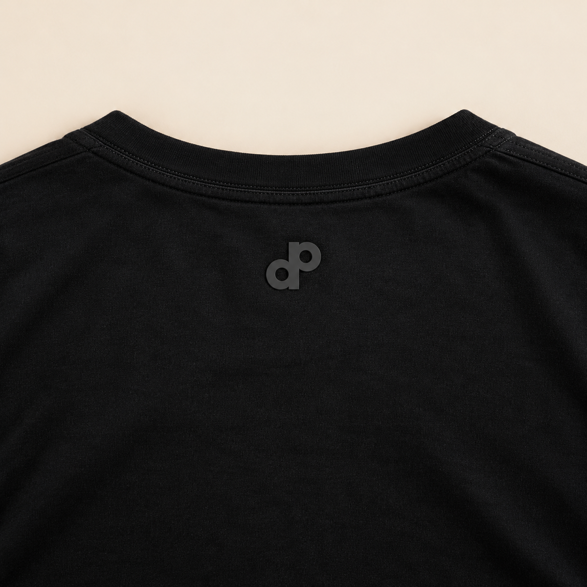 Rear neckline detail of the High Roller black shirt, with a small charcoal rubber DP logo centered below the back collar.